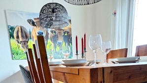Un comedor con una mesa de madera dispuesta para dos personas, una lámpara colgante tejida de color negro y un cuadro de vacas en la pared.