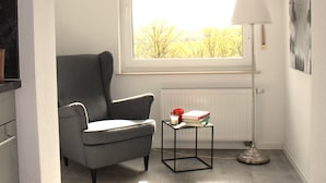Una habitación con un sillón gris, una mesita con libros y una vela, una lámpara de pie y una ventana con vistas a árboles.