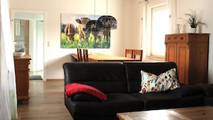 Una sala de estar con un sofá negro, un cojín rojo, una mesa de centro de madera, una mesa de comedor con sillas y un cuadro de vacas en la pared.