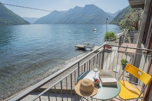 Un balcón con vista a un lago, una mesa con un libro y un sombrero, y una silla.