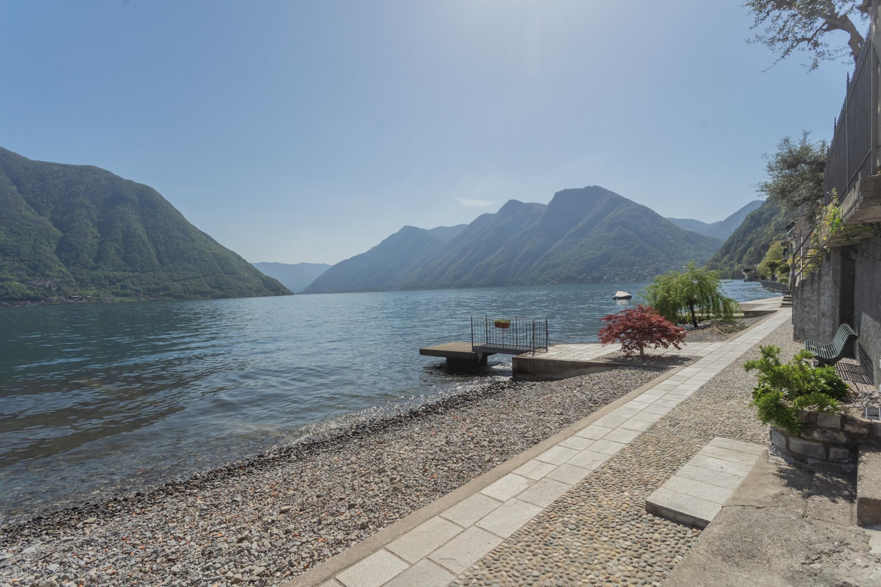 A lakeside walkway with pebbles, a bench, and a small garden area.