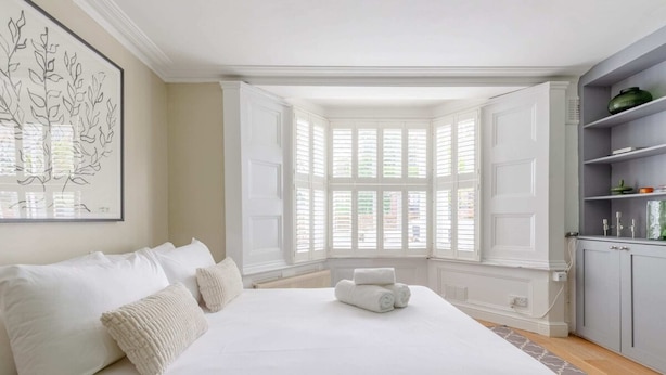Una cama grande con ropa blanca, vista al exterior a través de ventanales y un armario gris con estantes.