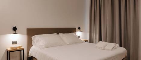 A neatly made bed with white linens, a bedside table with a book, and two wall-mounted lamps.
