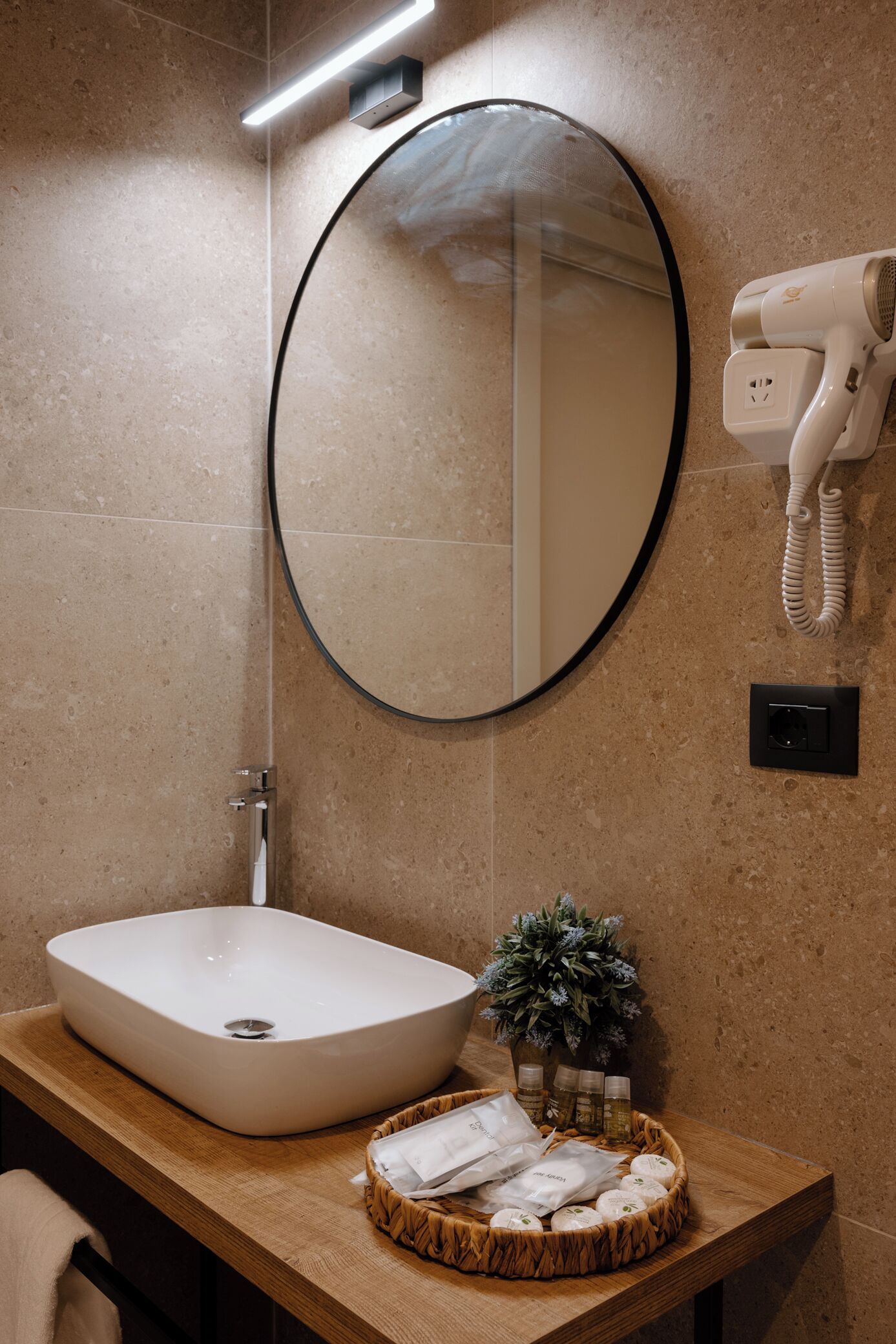 Un baño con un espejo ovalado grande, un lavabo blanco, un secador de pelo montado en la pared y una pequeña planta en maceta sobre la encimera.