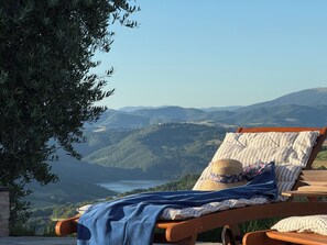 Una silla de madera para lounge con una toalla rayada y un sombrero de paja, con vistas a un paisaje montañoso con un lago.