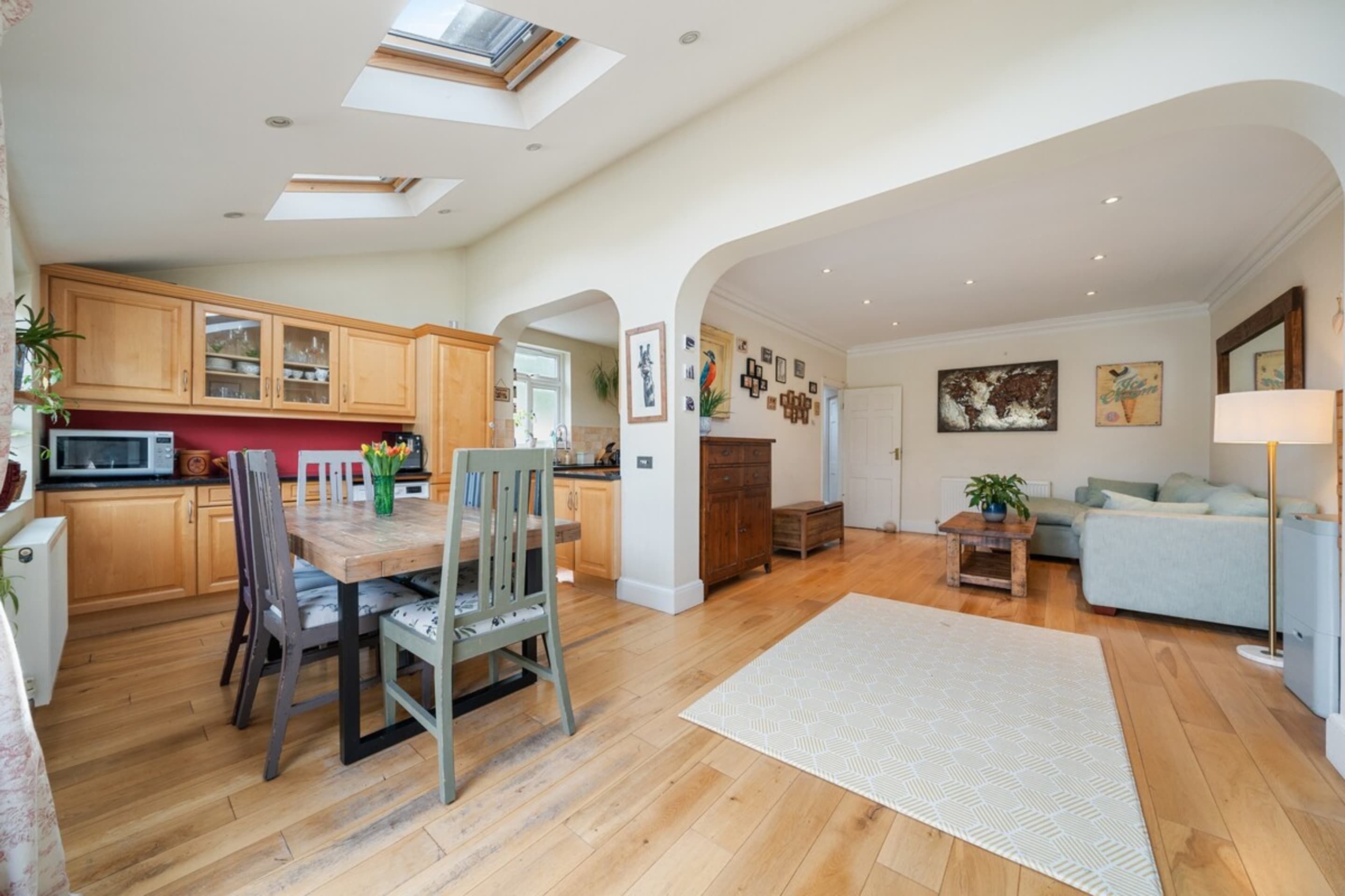 A spacious living area with a wooden dining table, chairs, and a sofa. There is a kitchen with wooden cabinets and a microwave. The room has a high ceiling with skylights and a large window.