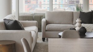 A modern living room with a sofa set, a coffee table, and a view of tall buildings through large windows.