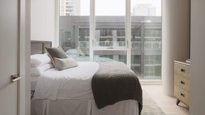 A bedroom with a bed, a dresser, and a large window with a view of buildings.