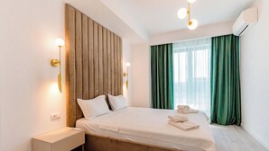 A modern hotel room with a wooden headboard, a bedside table with a lamp, a neatly made bed, and a large window with green curtains. - Wave 4 Residence (Navodari)