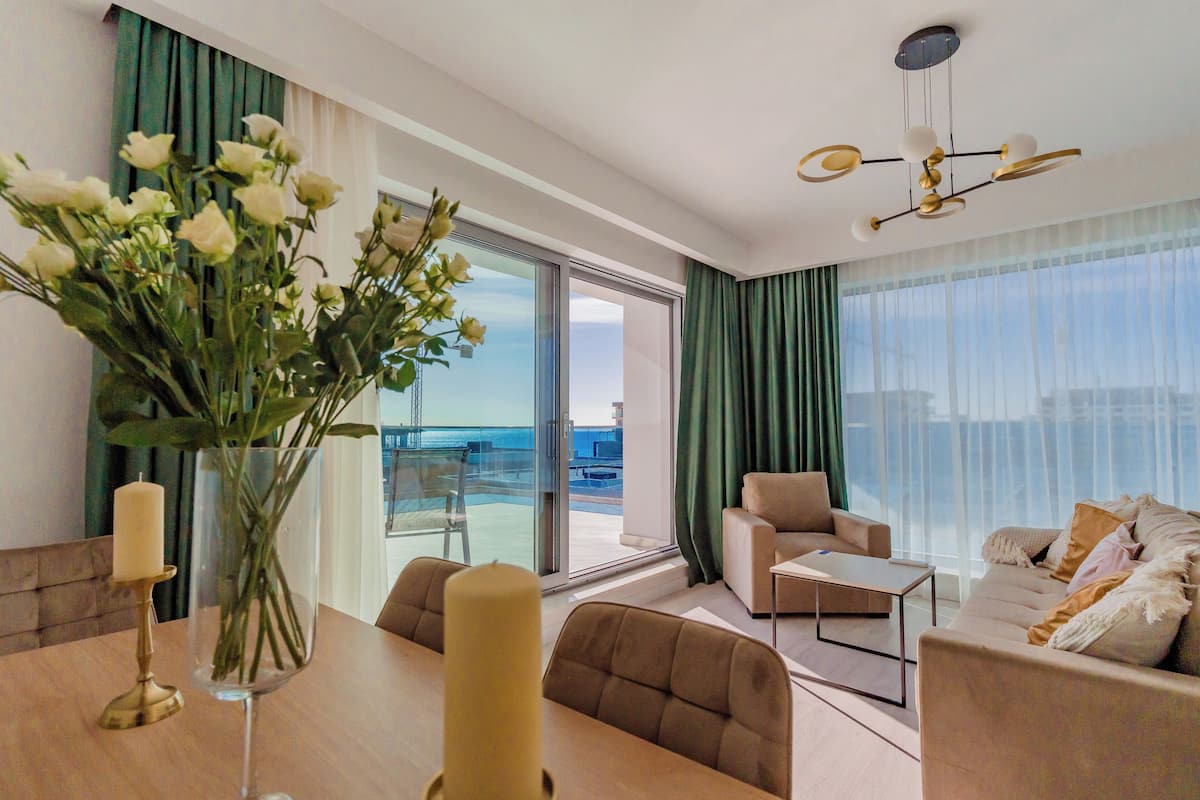 Una sala de estar moderna con un sofá, una mesa de centro y un comedor con una mesa y sillas. Hay un ventanal con cortinas, y un jarrón con flores sobre la mesa.