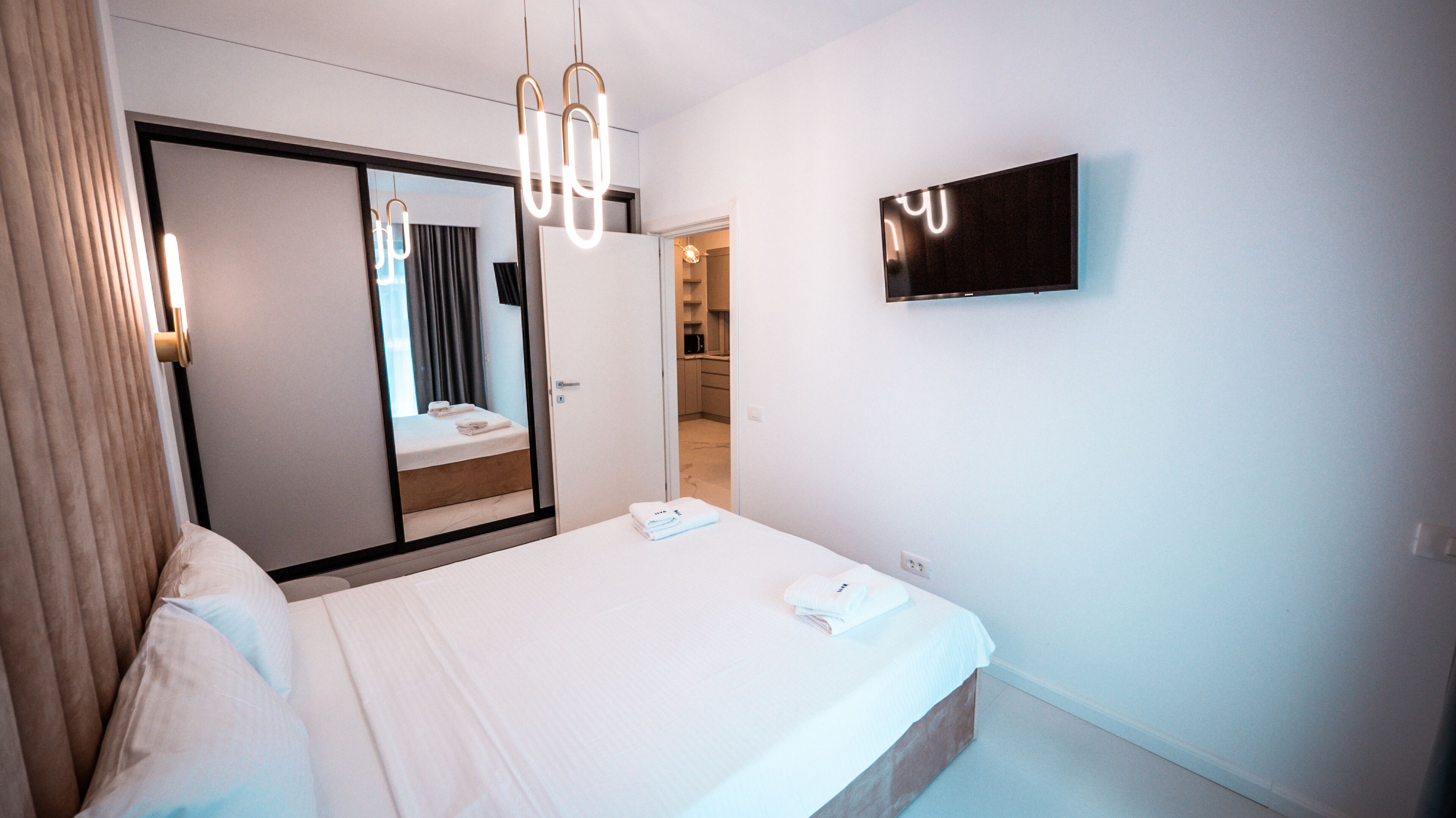 A modern hotel room with a large bed, a flat-screen TV mounted on the wall, and a mirror reflecting the room.