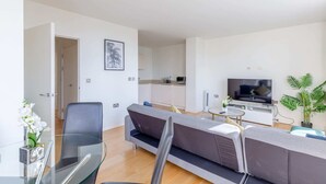 Un salon moderne avec un canapé, une table en verre et un téléviseur. On aperçoit une cuisine au fond.