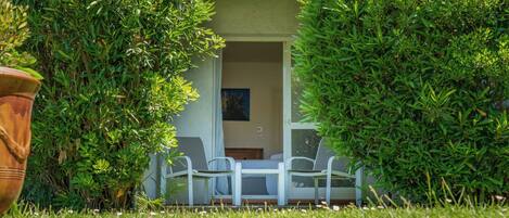 A small, enclosed outdoor seating area with a table and chairs, surrounded by lush greenery and a potted plant.