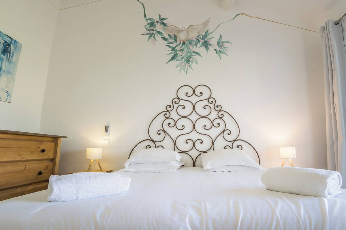 Un lit bien fait, avec du linge de lit blanc, une tête de lit décorative en métal, des lampes de chevet et une commode en bois.