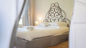 Una cama bien hecha con sábanas y almohadas blancas, un cabecero de metal con diseños intrincados y una lámpara de noche sobre una mesita de madera.