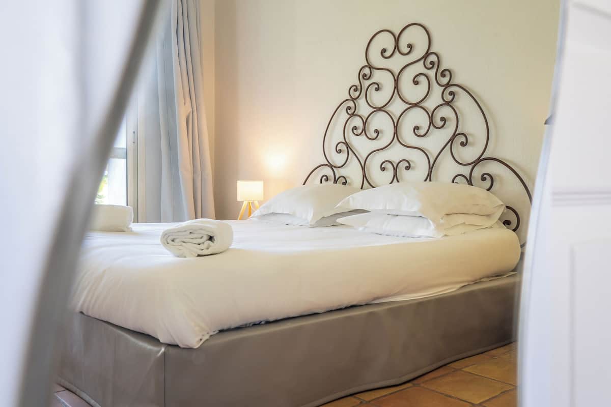Un lit bien fait, avec du linge de lit blanc et des oreillers, une tête de lit en métal ornée de motifs complexes, et une lampe de chevet posée sur une table de nuit en bois.