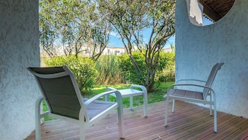 Un espacio al aire libre cubierto con dos sillas blancas y una mesa, con vista a un jardín y un edificio a lo lejos.