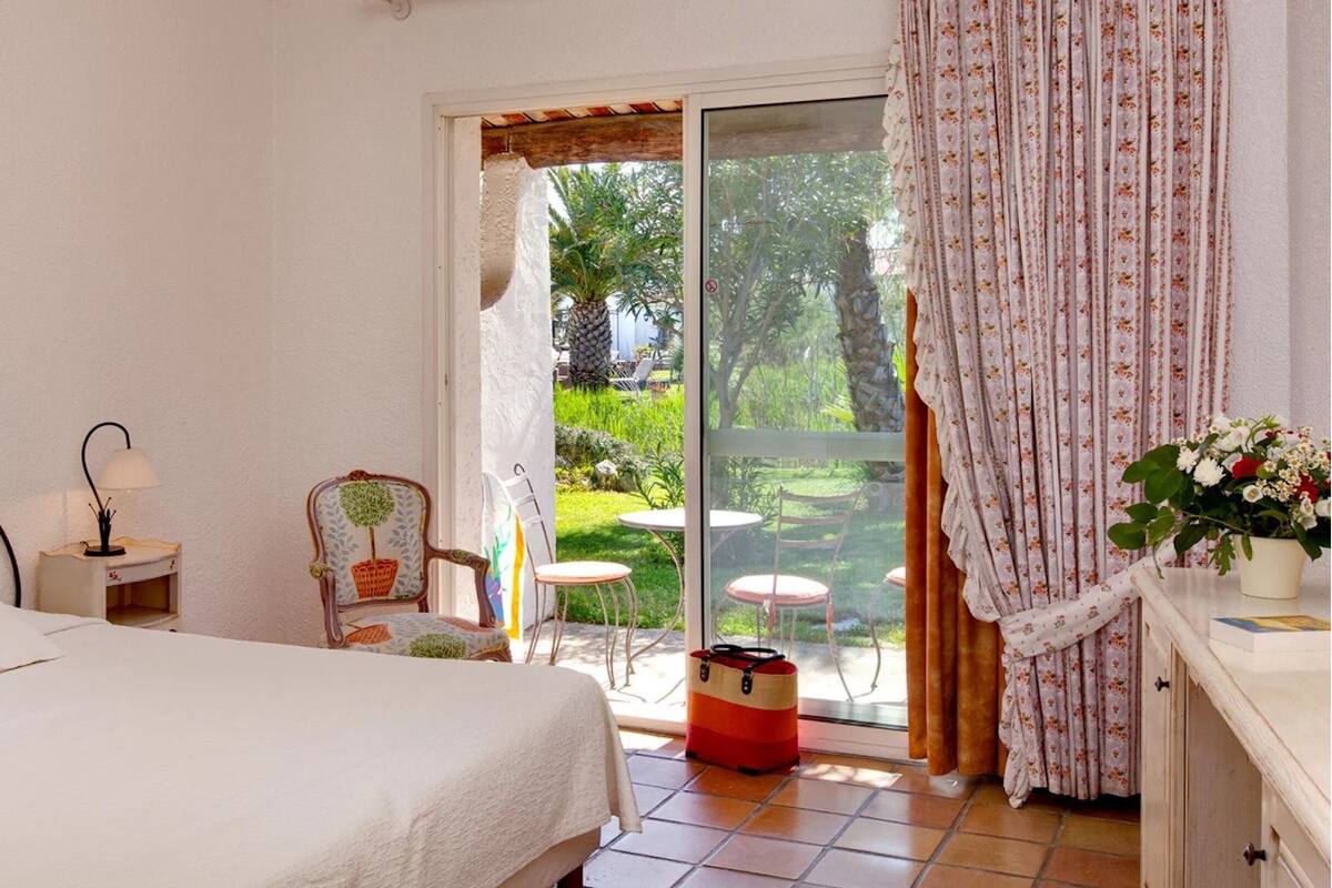 Une chambre à coucher comprenant un lit, une table de chevet, une chaise et une vue sur un jardin, accessible par une porte-fenêtre coulissante.