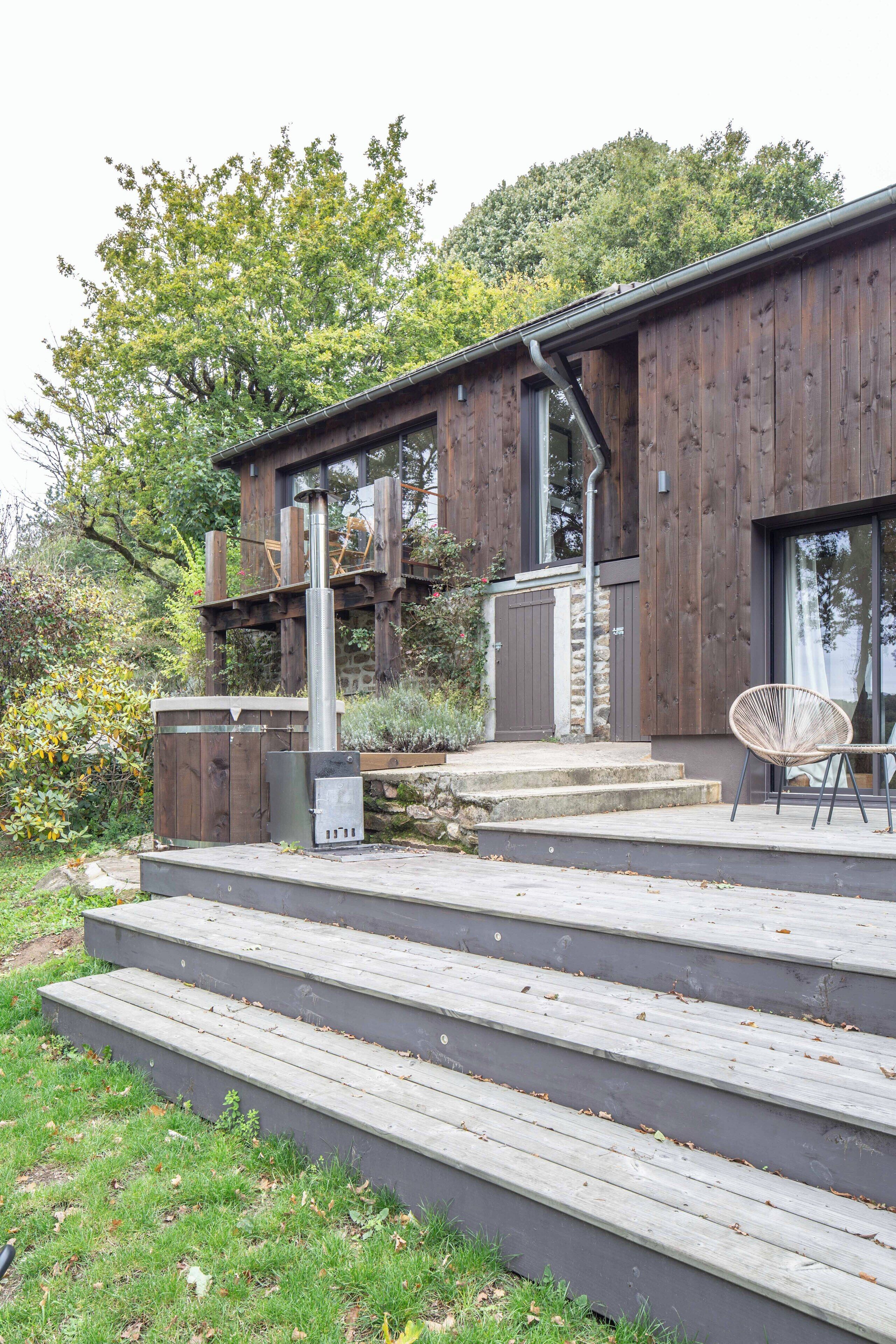 A modern wooden building with a stone foundation, a metal chimney, and a wooden deck with steps leading to a seating area with a chair.