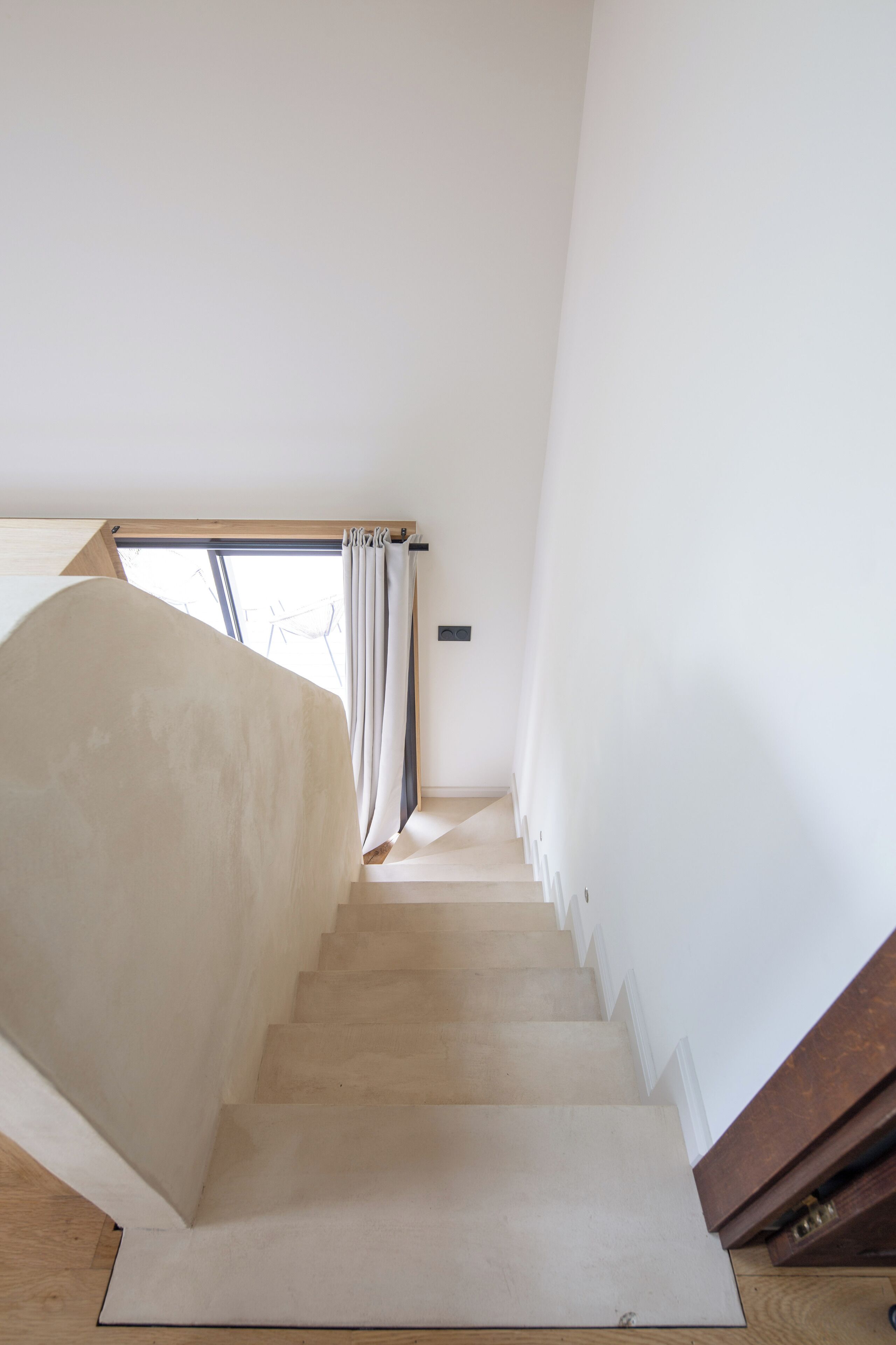 A staircase with a wooden handrail and a view of the outdoors through a window with curtains.