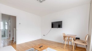 Un salon moderne avec un plancher en bois, un tapis gris, une petite table en bois avec une télécommande, un téléviseur fixé au mur et un coin repas avec une table et deux chaises.