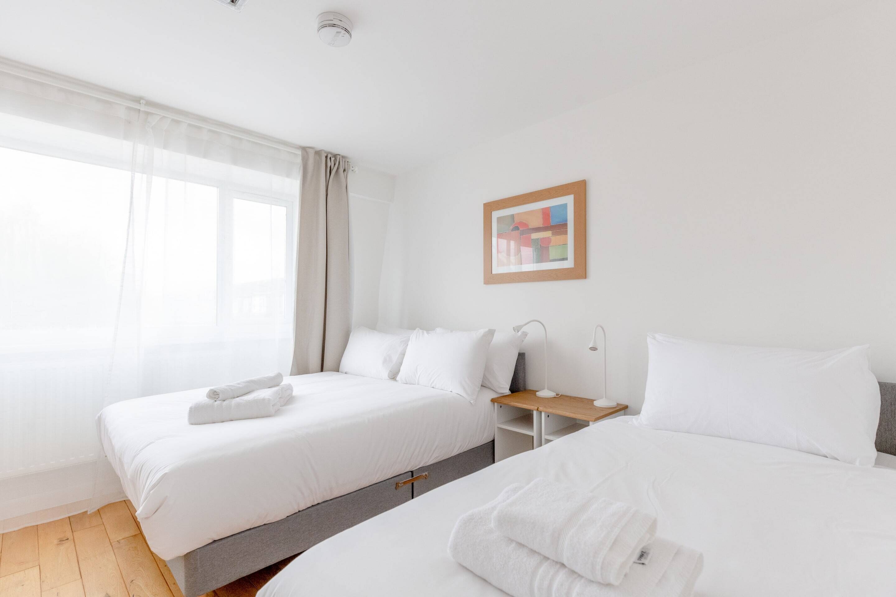 A hotel room with two beds, a bedside table, a lamp, and a window with sheer curtains.