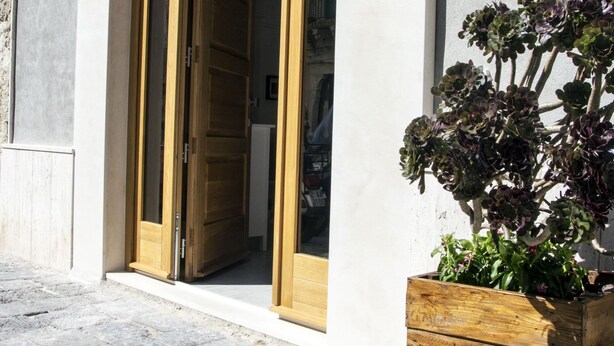 A building entrance with wooden doors, a potted plant, and a number '62' sign on the wall. - Casa Delle Mancarelle (Syracuse)
