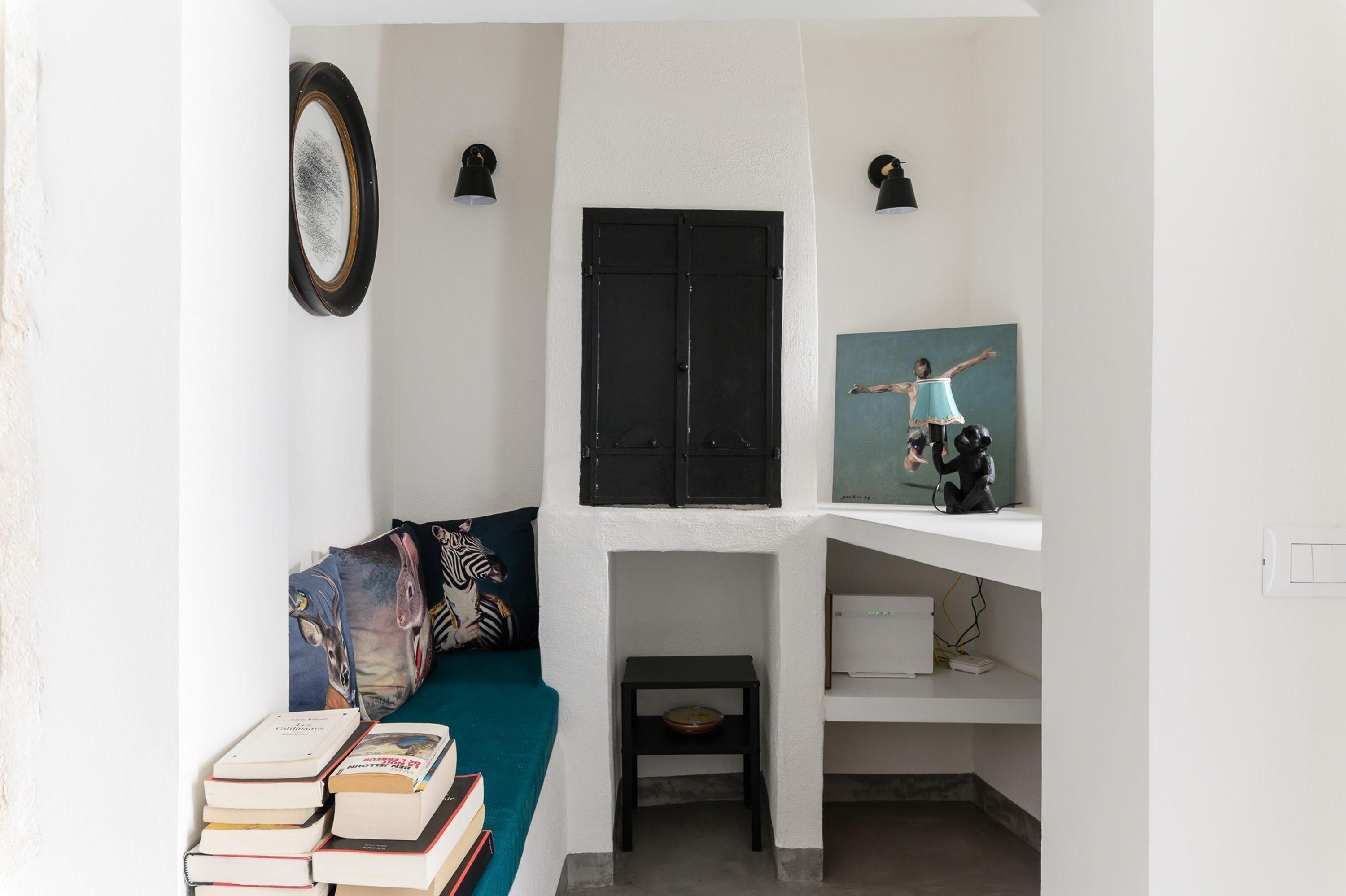 A compact living space with a teal sofa, a small table with books, a black-framed mirror, and a painting on the wall.