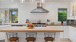A modern kitchen with white cabinetry, a central island with stools, a stainless steel range hood, and pendant lighting. - Gorgeous Pool and Sweeping Views (Sonoma)