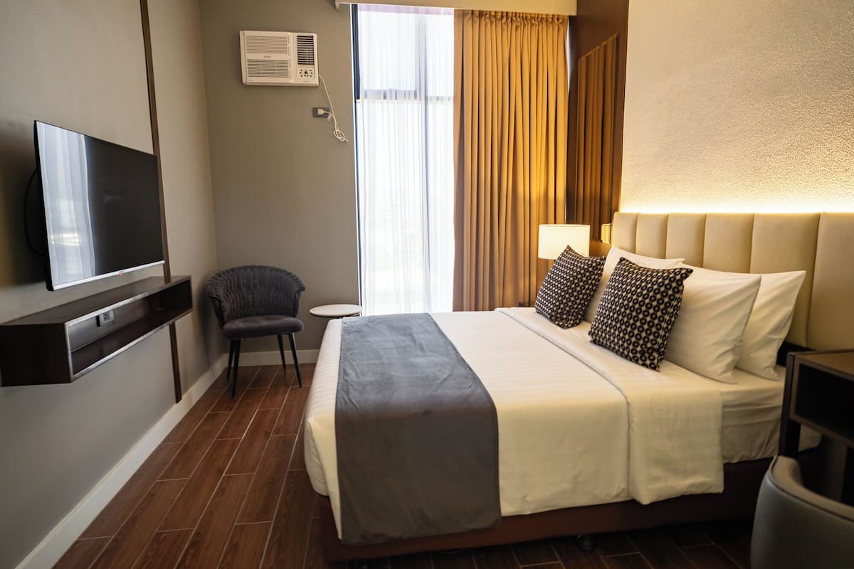 A hotel room with a bed, a TV, a chair, a small table, and a window with curtains.