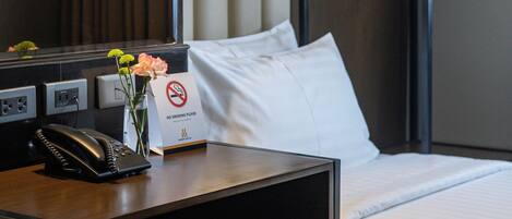 A hotel room with a bed, a desk with a phone, a vase with flowers, a no-smoking sign, and a hanging light fixture.