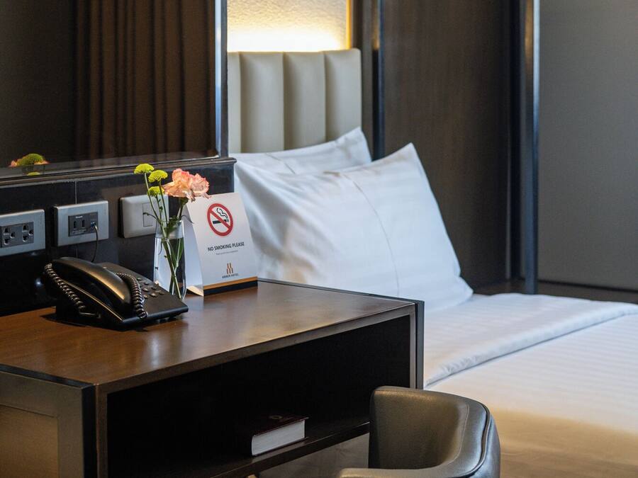 A hotel room with a bed, a desk with a phone, a vase with flowers, a no-smoking sign, and a hanging light fixture.