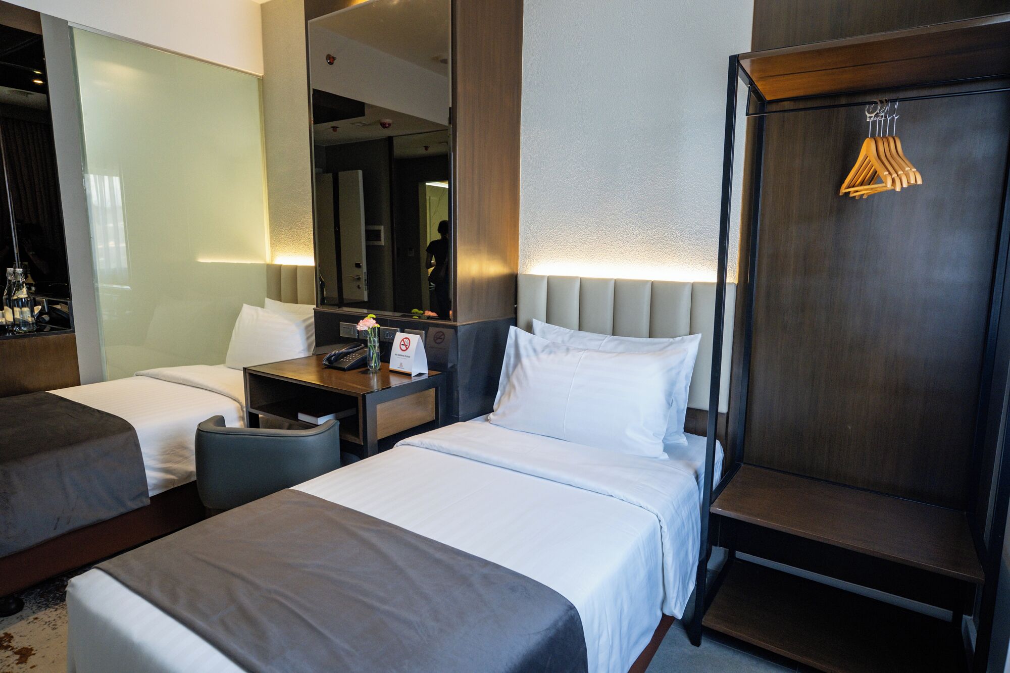 A hotel room with two beds, a desk with a phone and a vase of flowers, a mirror, and a wardrobe with hanging clothes.