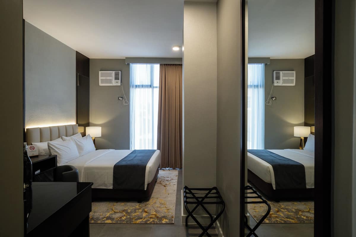 A hotel room with a bed, bedside tables, lamps, an air conditioning unit, and a view of the outside through a window with curtains.