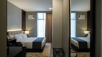 Habitación de hotel con cama, mesitas de noche, lámparas, unidad de aire acondicionado y vistas al exterior a través de una ventana con cortinas.
