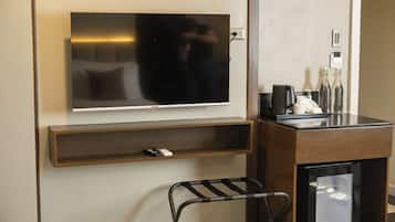 A hotel room with a flat-screen TV mounted on the wall, a small shelf below it, a coffee machine, a telephone, and a mini-fridge.