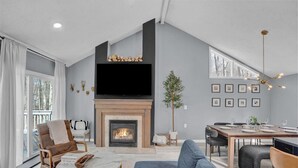 A modern living room with a fireplace, a dining area, and a large window overlooking a deck. - Pocono Blue | Paddleboards, Spa, Indoor Jacuzzi, GameRoom, Lake, EV (Clifton Township)