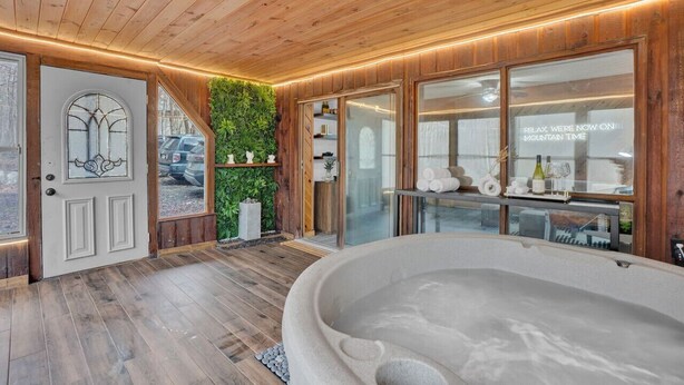 A spa room with a wooden ceiling, a large bathtub, and a glass door with a view of the outside. - Pocono Blue | Paddleboards, Spa, Indoor Jacuzzi, GameRoom, Lake, EV (Clifton Township)