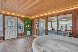 Une salle de spa avec un plafond en bois, une grande baignoire et une porte en verre donnant sur un espace de détente agrémenté d’étagères et d’un mur végétal.