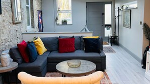 Un salon moderne avec un canapé d’angle, une table basse ronde et une petite table d’appoint. Une lucarne se trouve au-dessus du canapé, et une fenêtre offre une vue sur les arbres.