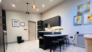 Une cuisine moderne avec des meubles de couleur sombre, un comptoir en marbre et un petit îlot central avec des tabourets. Des tableaux encadrés ornent le mur, et une porte donne accès à une autre pièce.