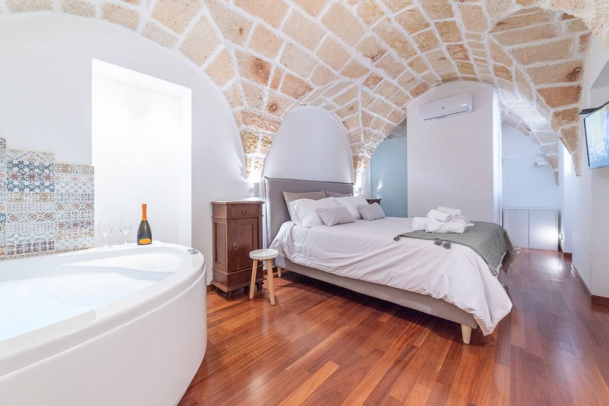 Een slaapkamer met een stenen plafond, een groot bed, een houten vloer, een klein krukje en een bad.