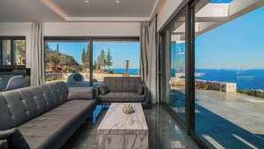 A modern living room with a sofa, armchair, and coffee table, large windows with a view of the sea and trees. - Nikolakos Villa - Pirvate Pool (Zakynthos)