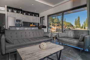 A modern living room with grey sofas, a marble coffee table, and a kitchen area with a 'ENJOY' sign.