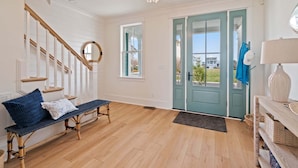 Een ruime kamer met houten vloer, een bank met kussens, een trap met witte leuningen en een blauwgroene deur naar buiten.
