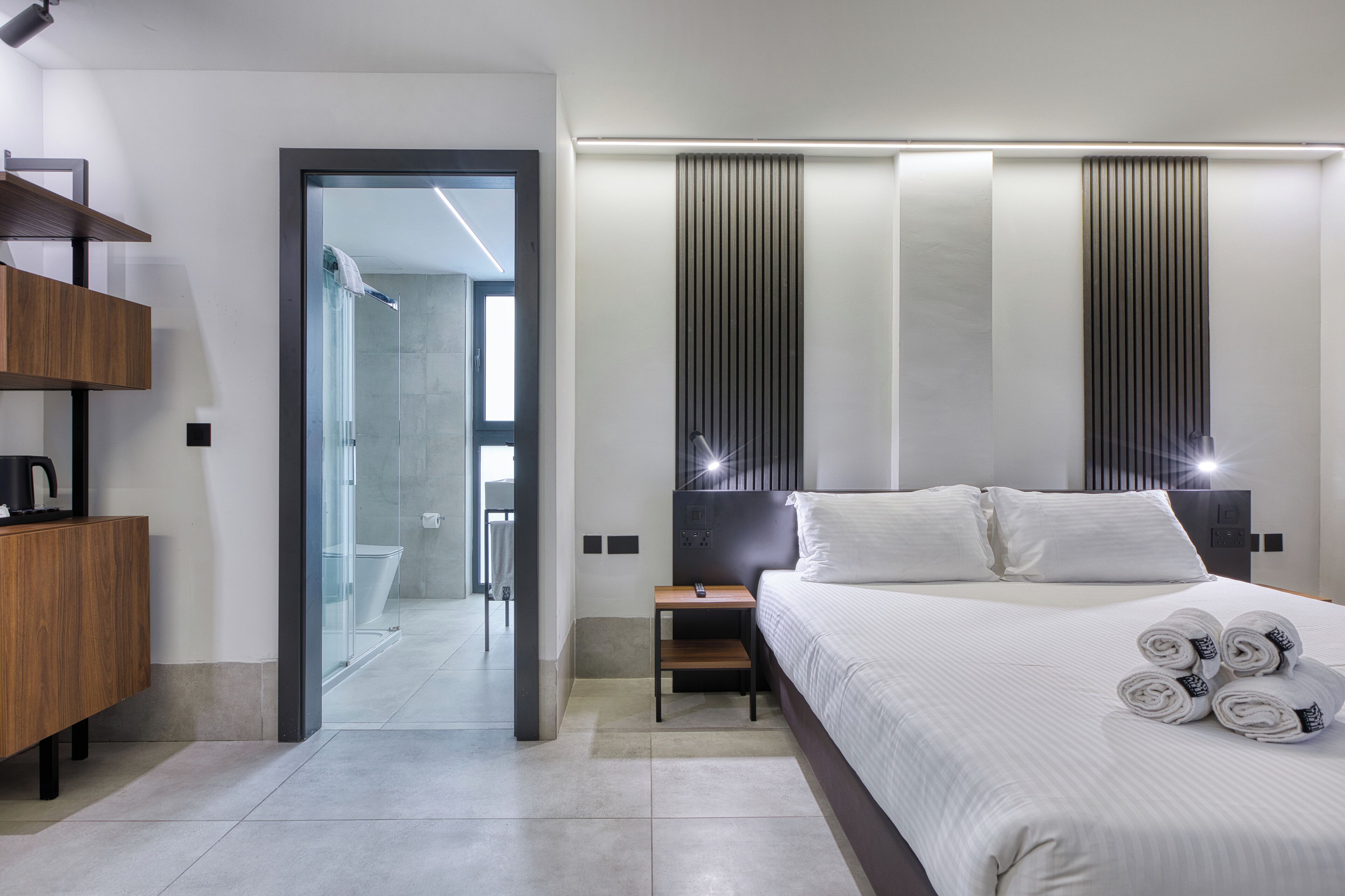 A modern hotel room with a large bed, a wooden side table, a nightstand, and a glass shower enclosure.