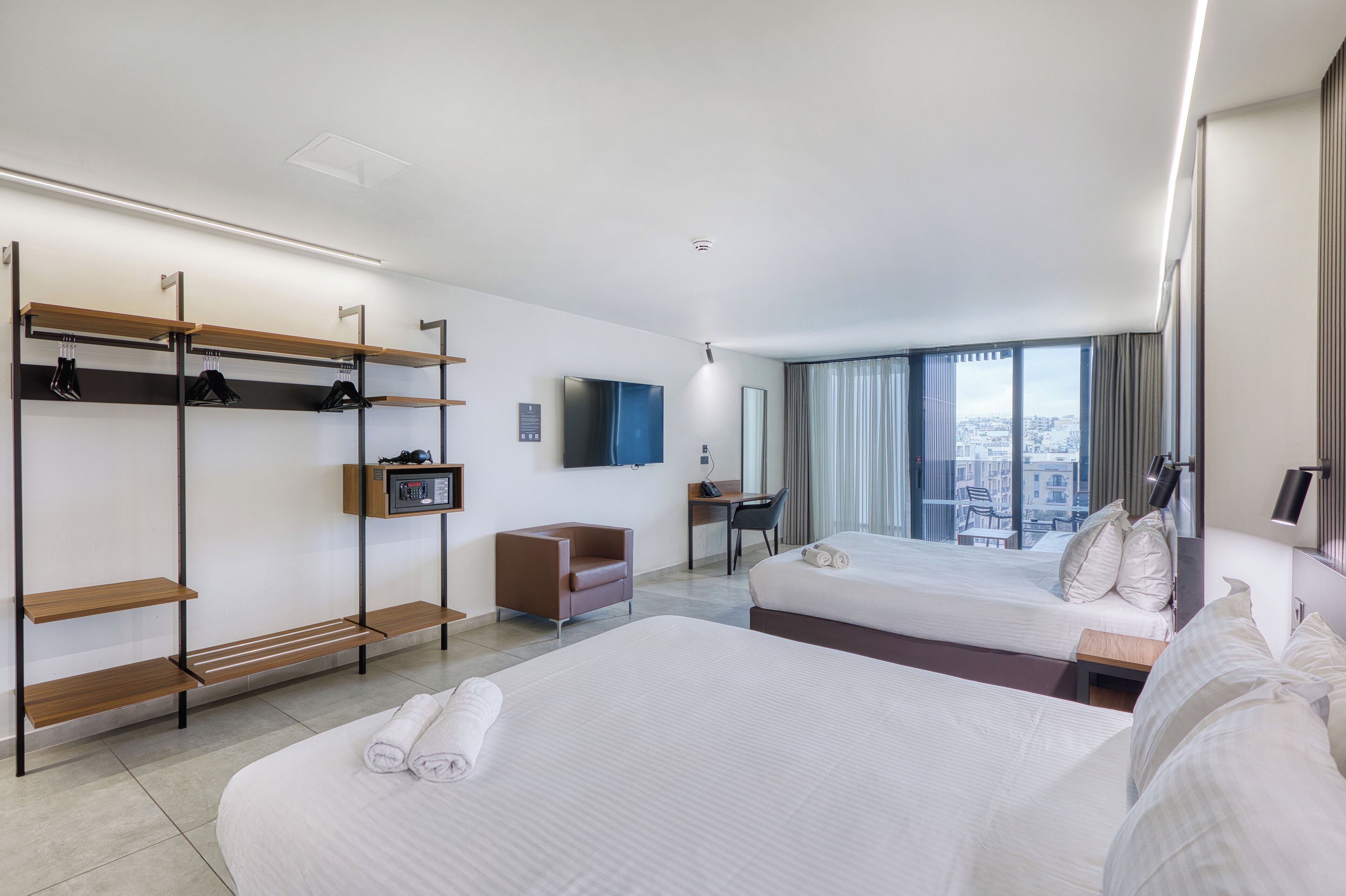Ein Hotelzimmer mit zwei Betten, einem Schreibtisch, einem Stuhl, einem Fernseher und Blick auf die Stadt.