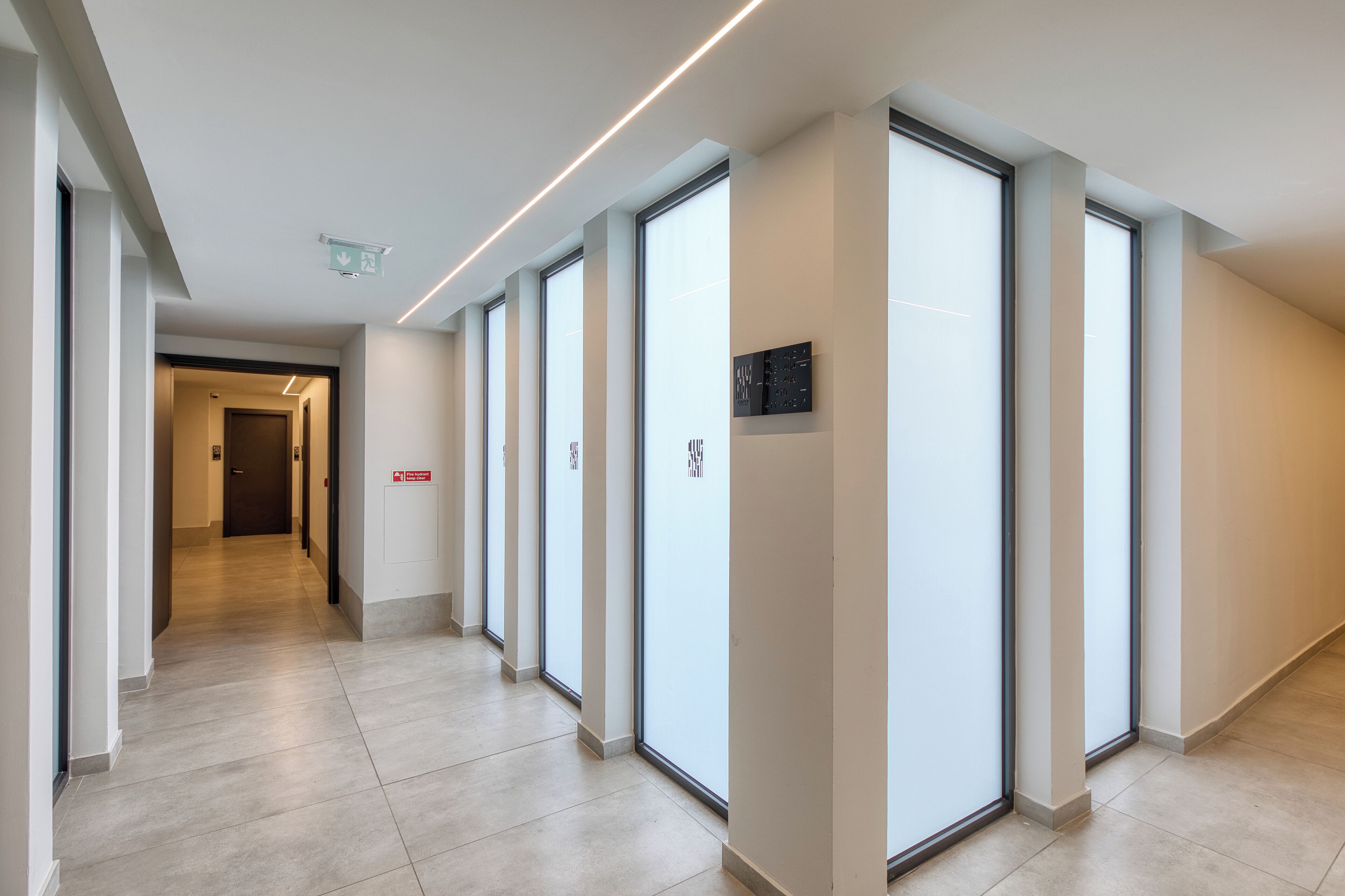 Un couloir moderne avec des portes en verre, un luminaire au plafond et un panneau indiquant la sortie de secours en cas d’incendie.