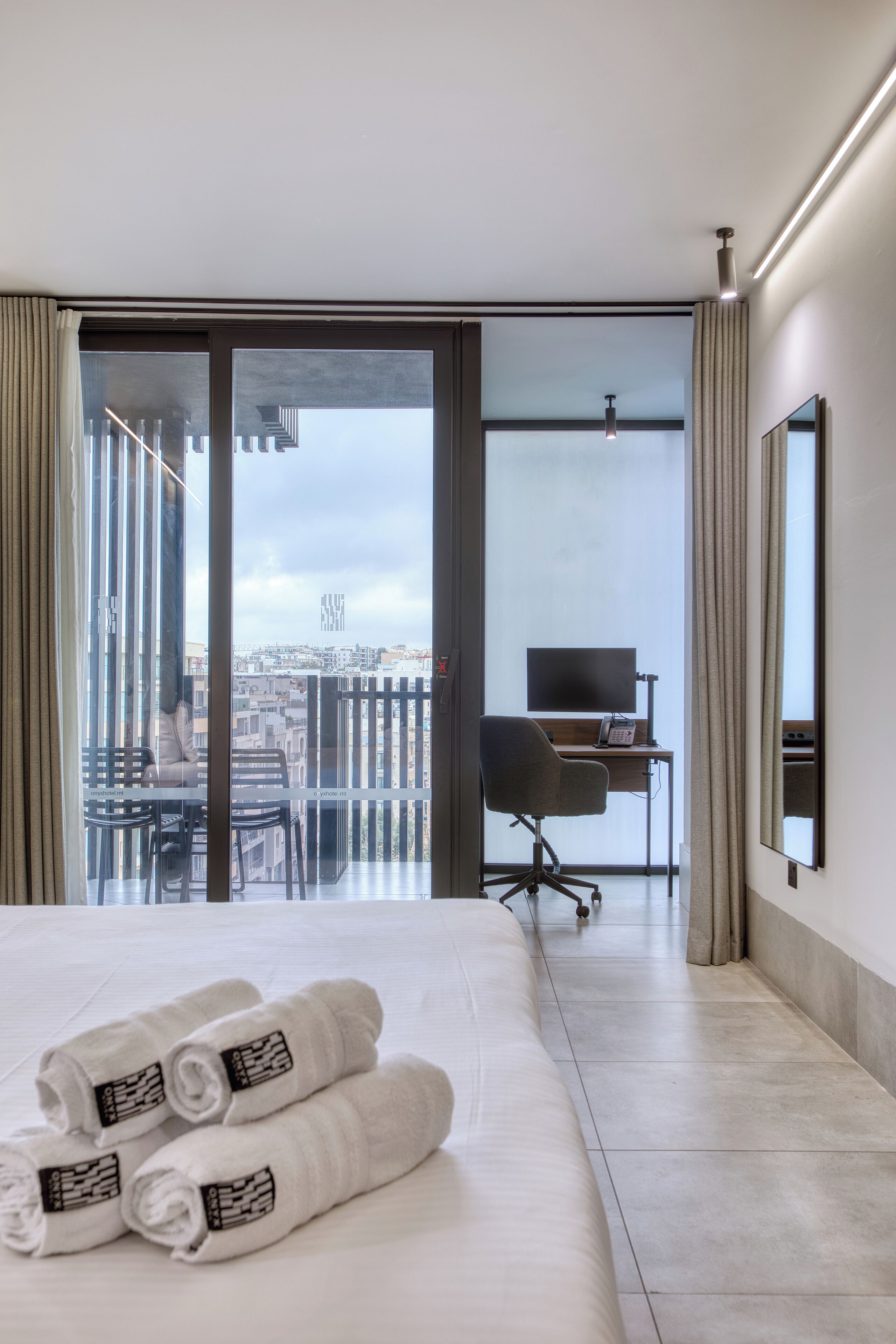 A hotel room with a bed, a desk with a computer, a chair, and a view of the city through large windows.