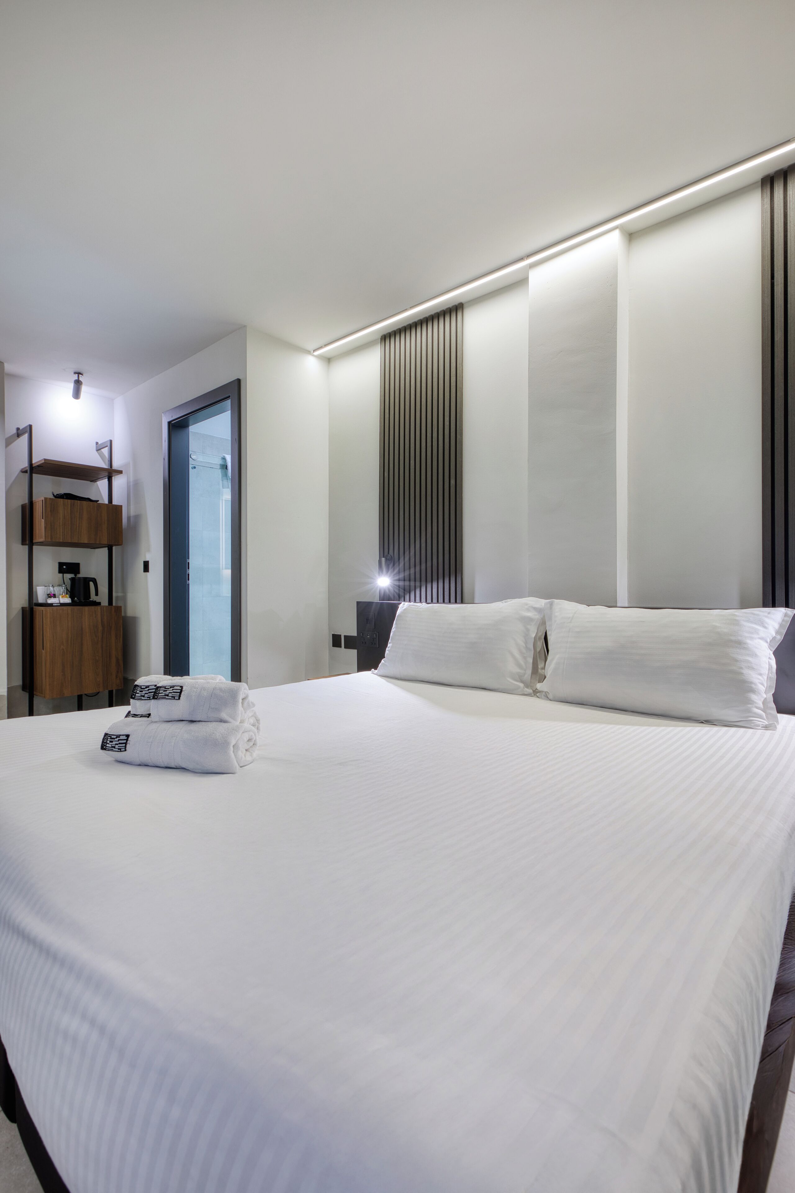 A modern hotel room with a large bed, a bedside table, a wardrobe, and a bathroom visible through a glass door.